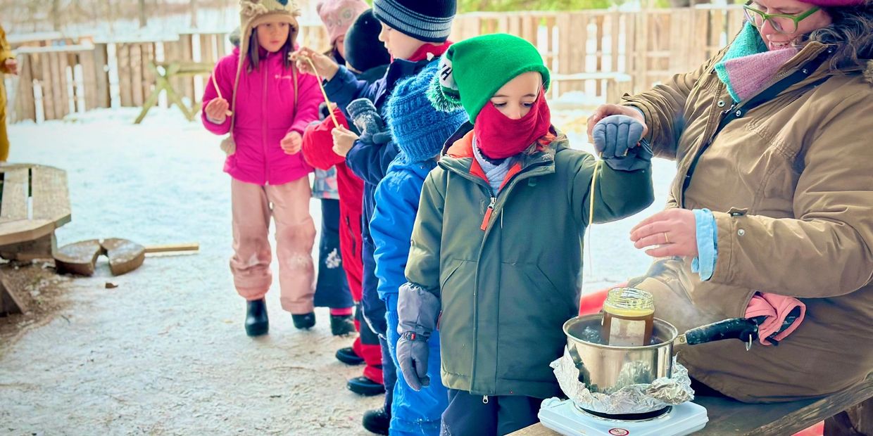 Children in winter clothes making maple taffy with an adult outdoors.
