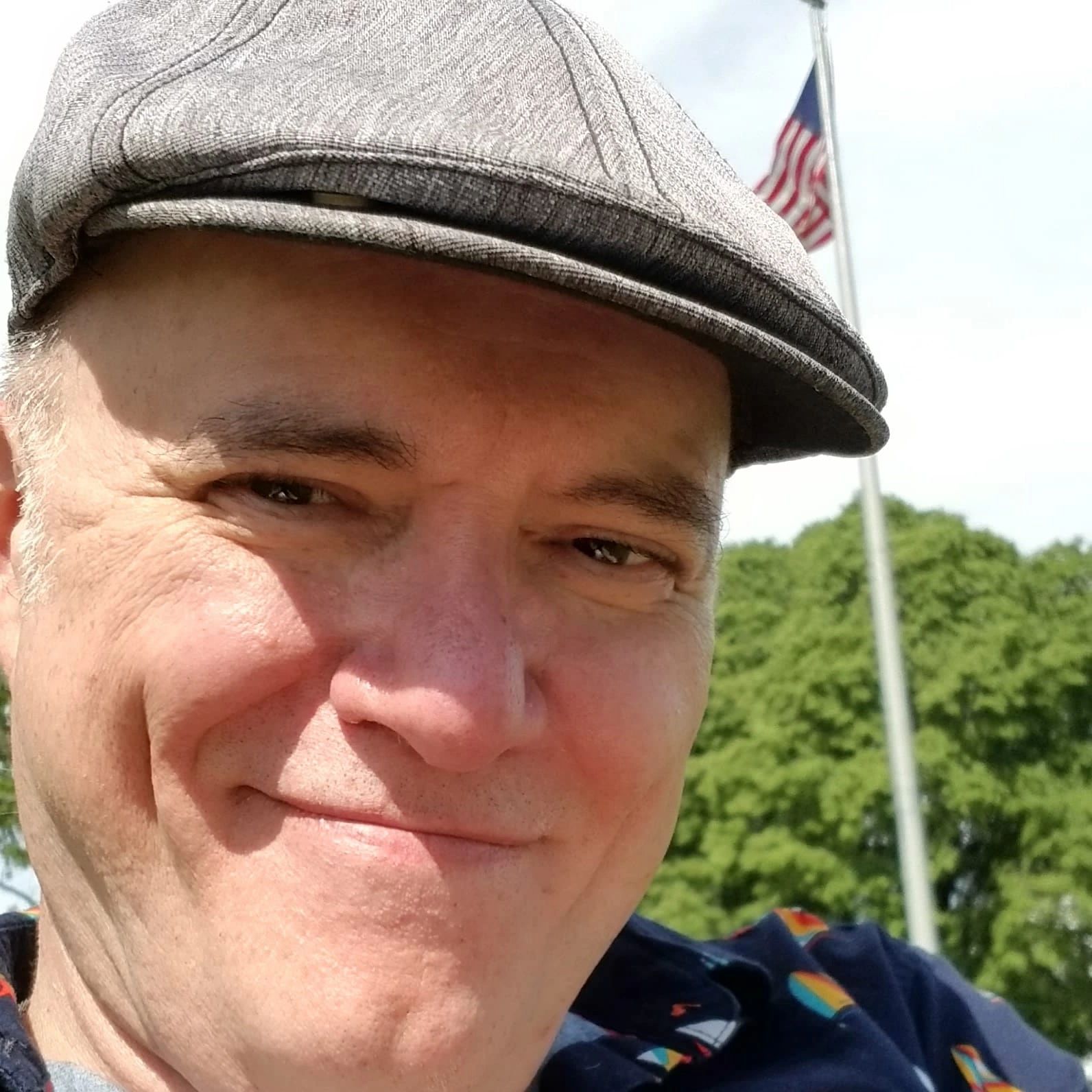 Close-up of a smiling man wearing a flat cap outdoors with a flag and trees in the background.
