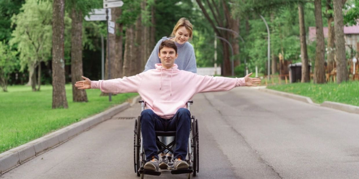 A young man in a wheelchair being pushed by a smiling woman on a tree-lined path.