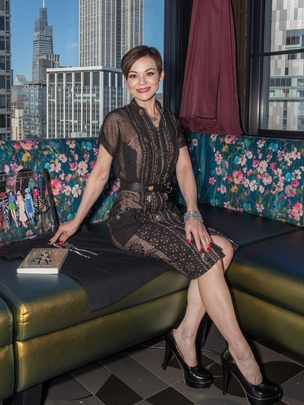 Woman in a stylish black dress sitting on a floral couch with city buildings in the background.