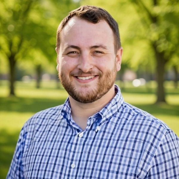 Smiling man in a blue plaid shirt standing outdoors with green trees in the background.