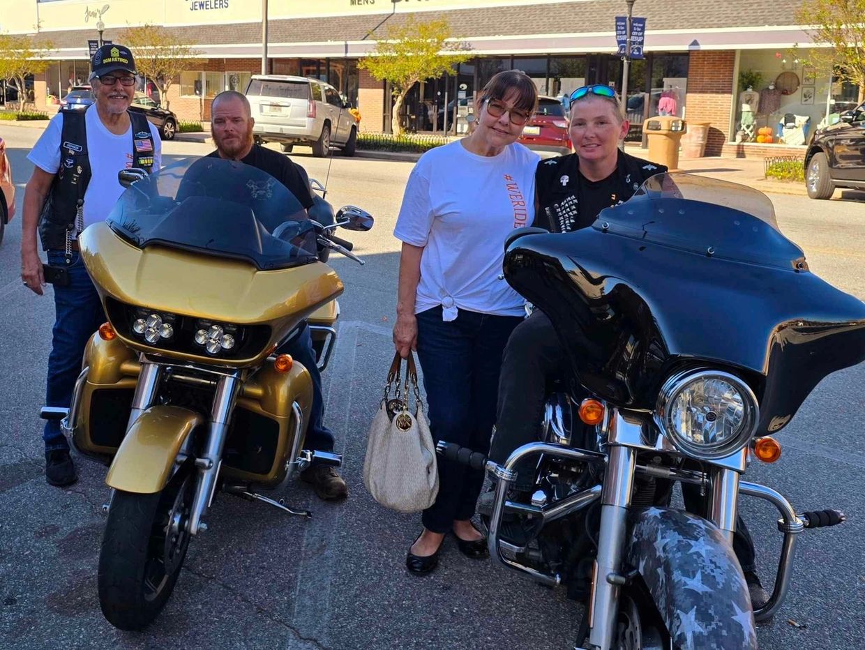 Four people posing with two motorcycles on a sunny day in a downtown area.