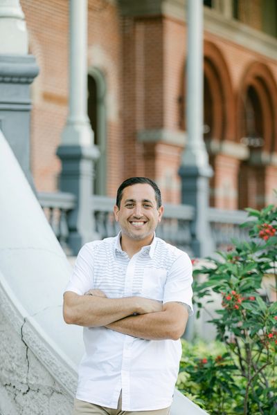 A smiling man in a white shirt stands with arms crossed by a garden and brick building.