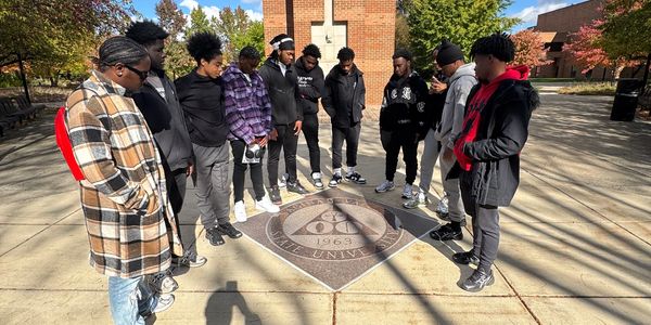 A group of young men standing in a circle around a university emblem on a sunny day.
