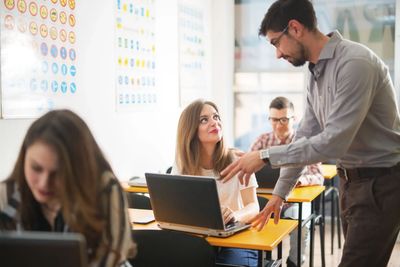 Teacher assisting a student working on a laptop in a classroom.