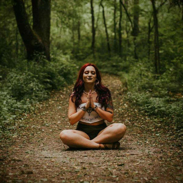Woman meditating in a serene forest path with eyes closed and hands in prayer.