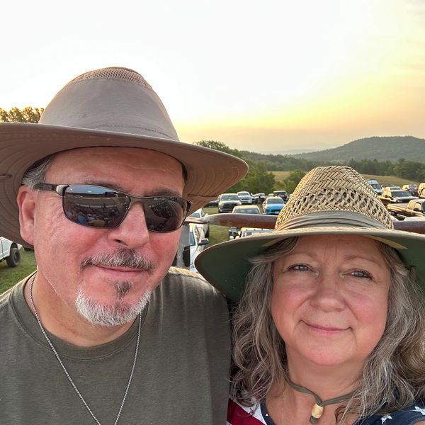Couple wearing hats outdoors with cars and hills in background.