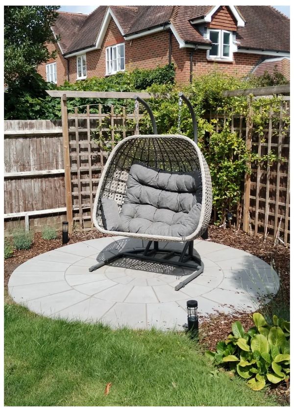 Circular stone patio with an egg chair resting on top. Weathered fence & trellis in Newdigate.