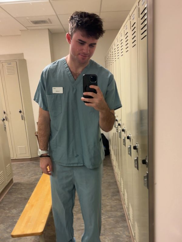 Young man in medical scrubs taking a mirror selfie in a locker room.