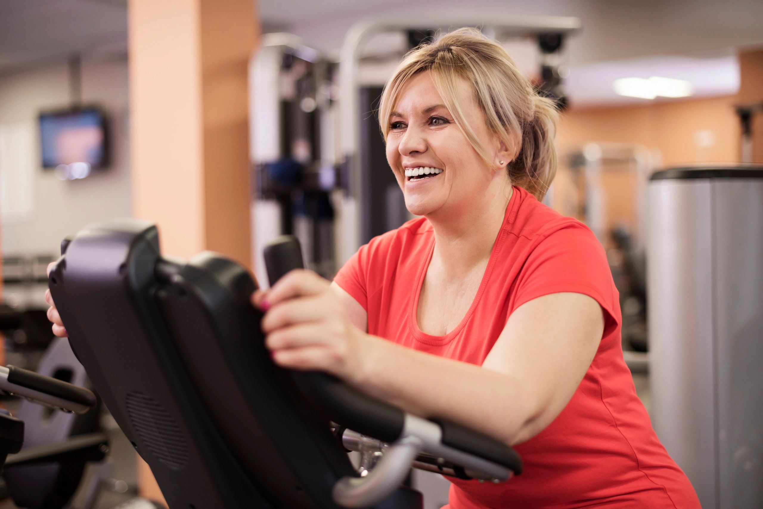 Smiling woman exercising on a stationary bike in a gym.