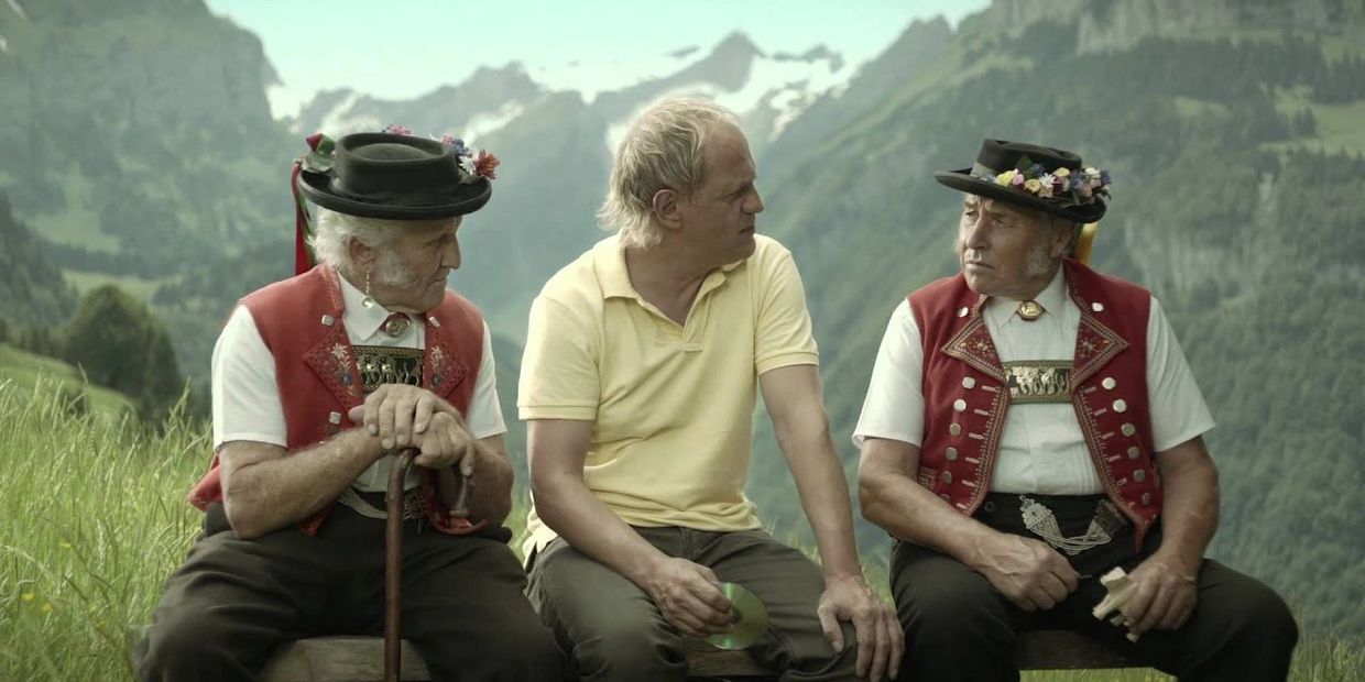 Der Schauspieler Uwe Ochsenknecht sitzt auf einer Bank zwischen zwei Sennen in Appenzeller Tracht.