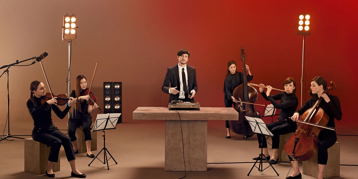 Ein gut gekleideter DJ umringt von fünf Musikerinnen, die klassische Instrumente spielen