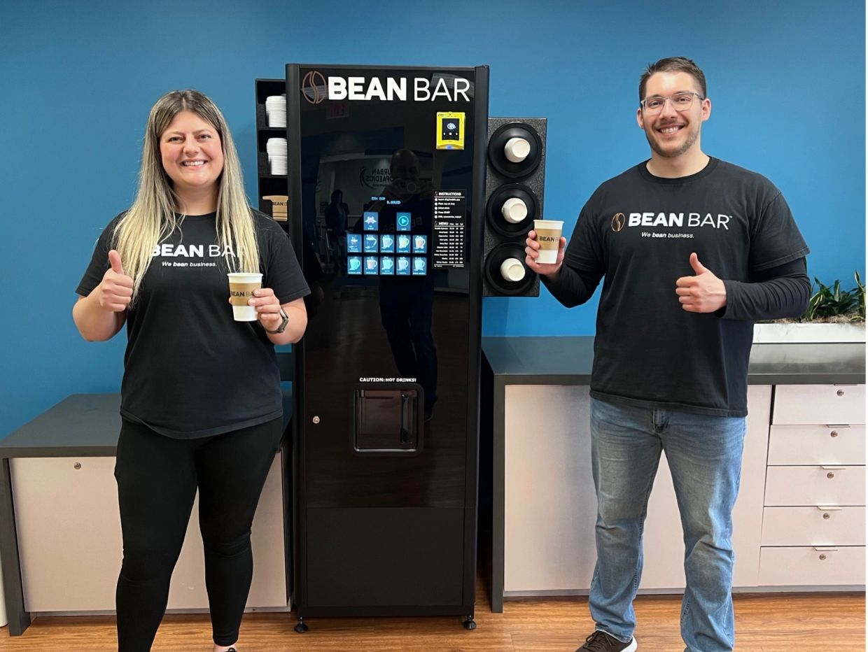 Two people holding coffee cups by a BEAN BAR machine giving thumbs up.