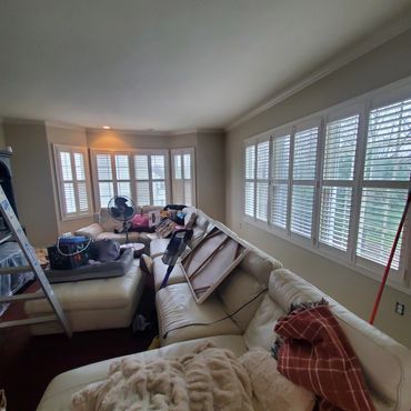 Living room with white sectional sofa, large windows with shutters, and various items scattered around.