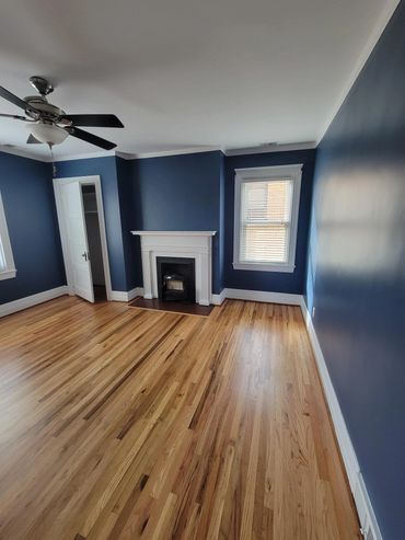Empty room with blue walls, hardwood floor, ceiling fan, and a white fireplace.
