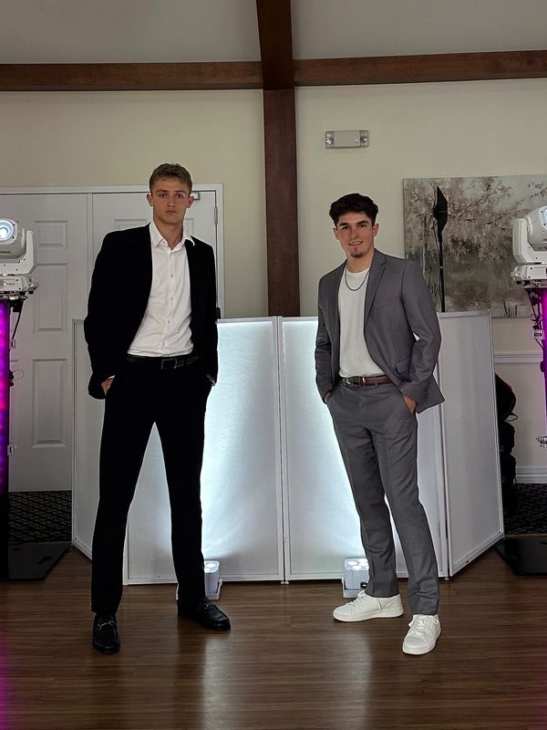 Two young men dressed in suits posing indoors with purple lights behind them.