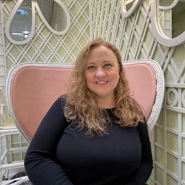 Woman with curly hair sitting on a pink cushioned chair with a decorative lattice background.