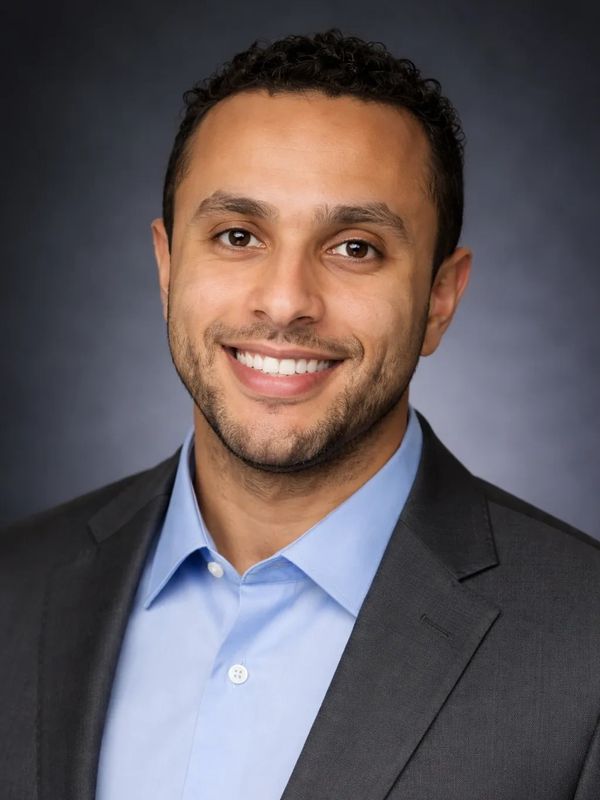 Smiling man in a suit and blue shirt against a dark background.