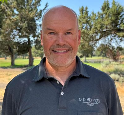 Smiling man in a black polo shirt outdoors with trees in the background.