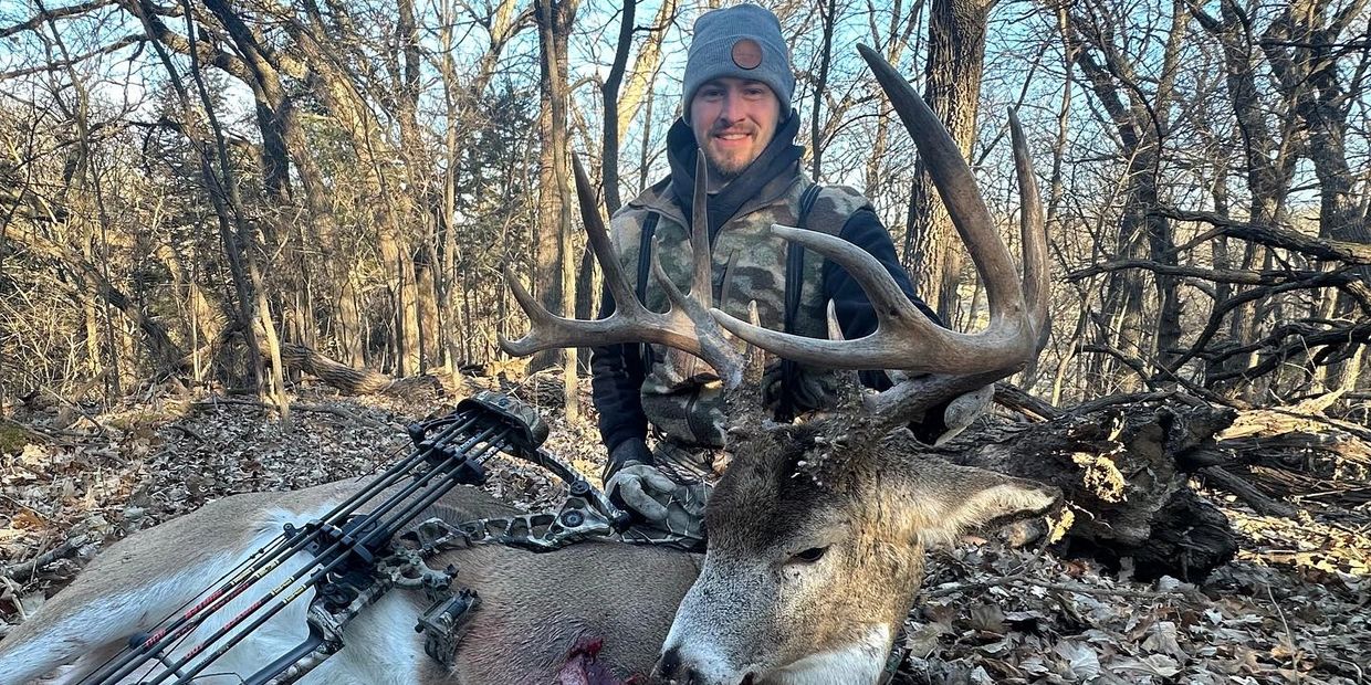 Hunter posing with a large deer and bow in a forest.