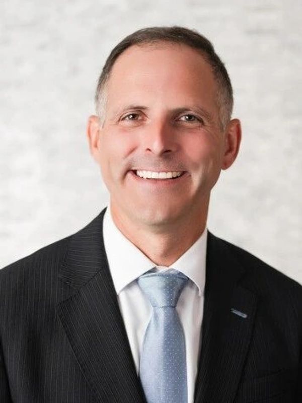 Smiling man in a suit and light blue tie against a soft background.