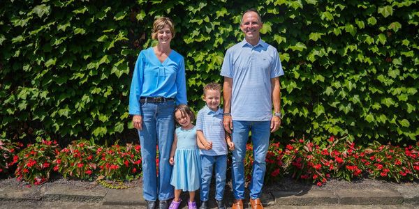 A family of four posing happily in front of a leafy green wall with red flowers.