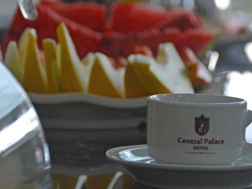Xícara de café com logo do hotel com frutas do café da manhã ao fundo.