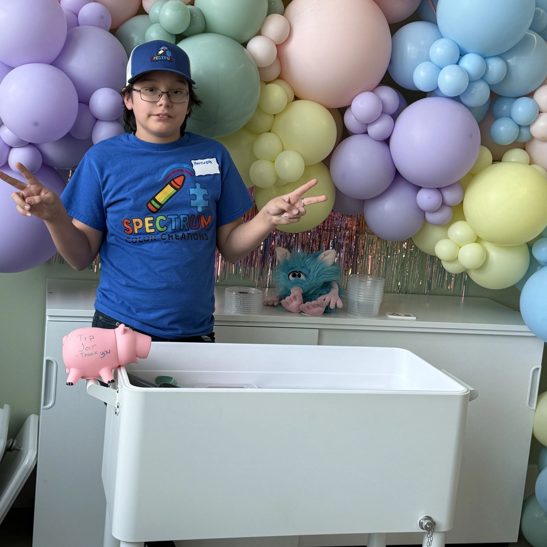 A boy in a blue Spectrum Color Creations shirt posing with peace signs in front of pastel balloons.