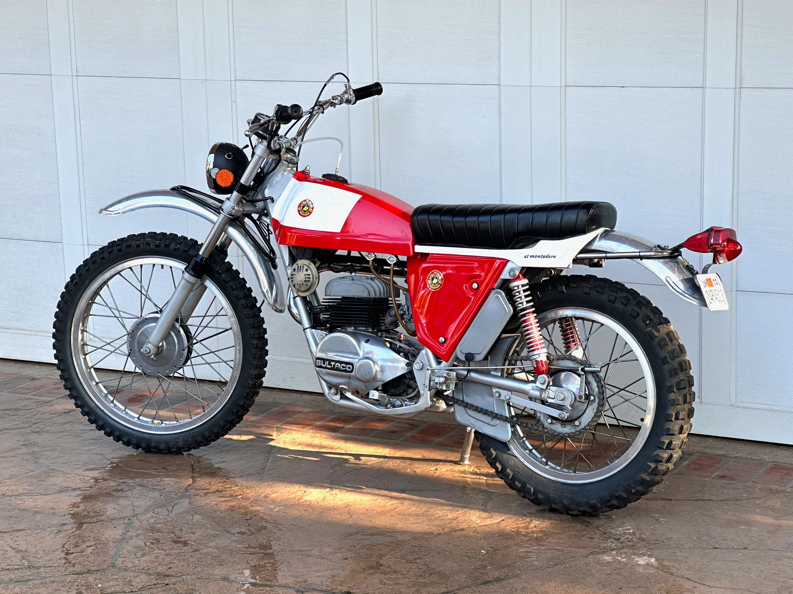 Classic red and white Bultaco El Montadero motorcycle parked outside a garage.
