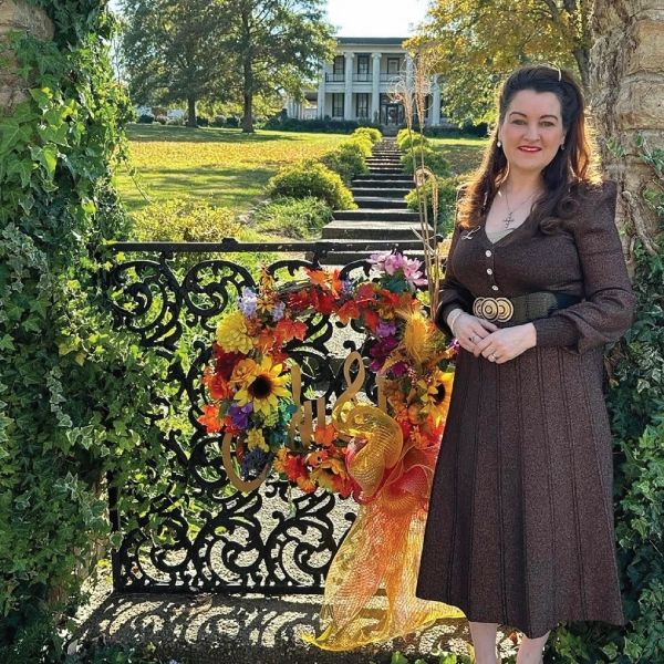 A woman in a brown dress stands by a decorative gate with a colorful wreath and a large house in the background.