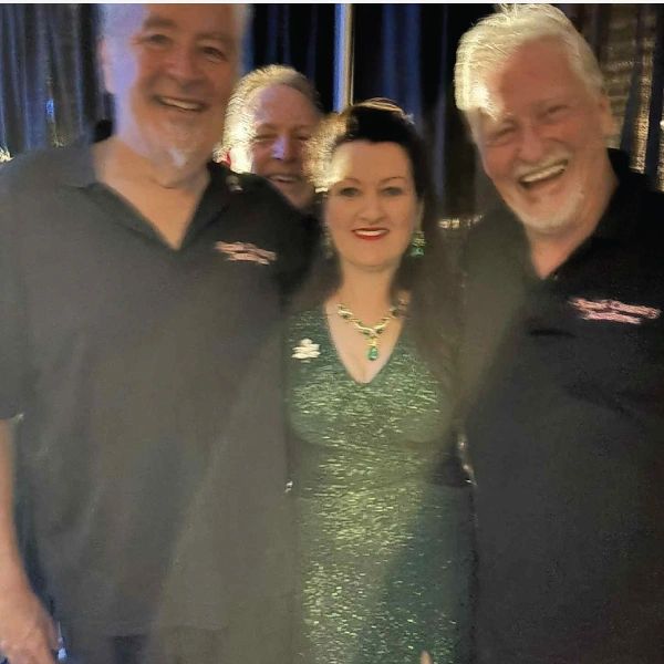 Group of four people smiling, three men in black shirts and one woman in a sparkling green dress.