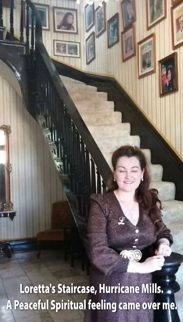 A woman stands smiling near a vintage staircase adorned with framed photos.