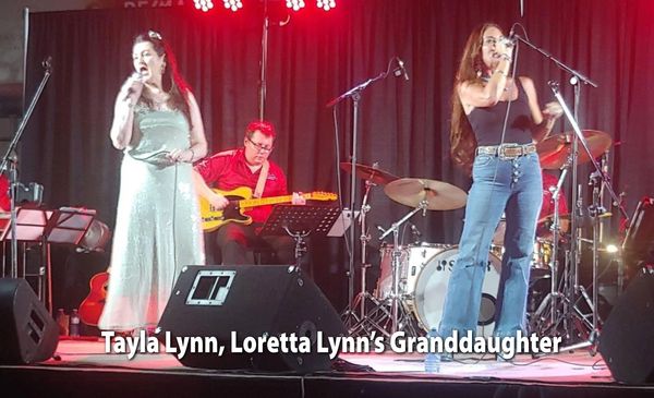Two women singing on stage with a guitarist and drummer in the background.