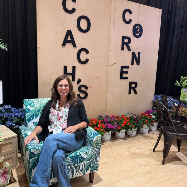 Woman sitting in a floral armchair next to a wall with "Coach's Corner" signage.