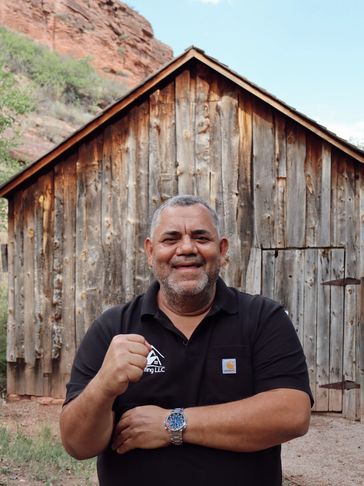 Smiling man in black shirt stands in front of a rustic wooden cabin outdoors.