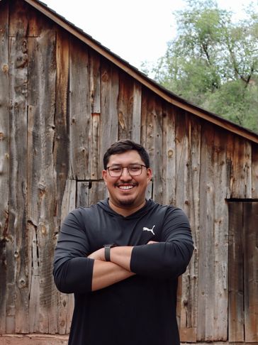 Smiling man with glasses and crossed arms in front of rustic wooden barn.
