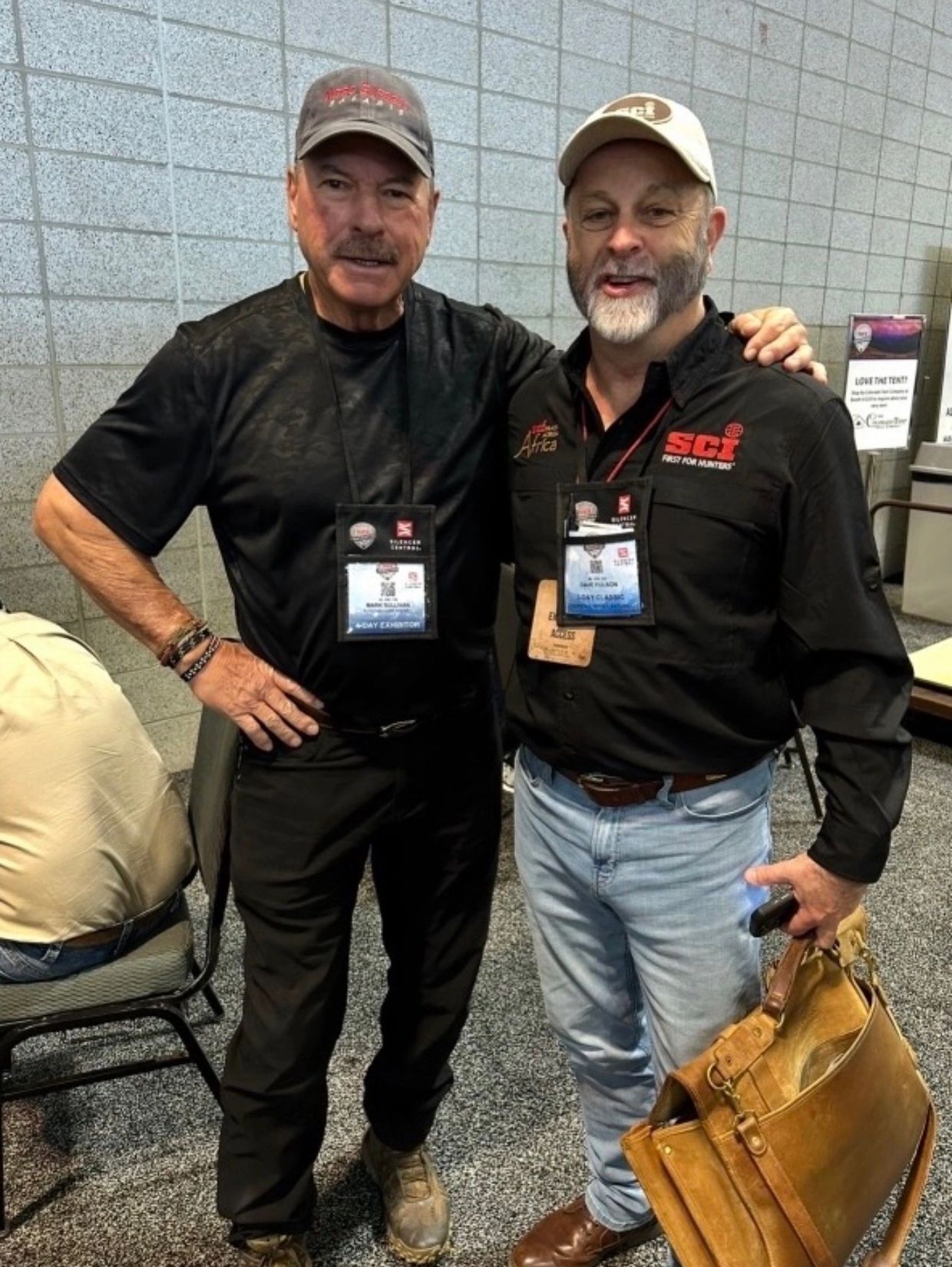 Two men posing together at an event, wearing badges and casual outdoor attire.