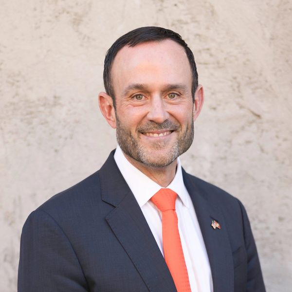 Man in a dark suit and orange tie smiling against a plain background.
