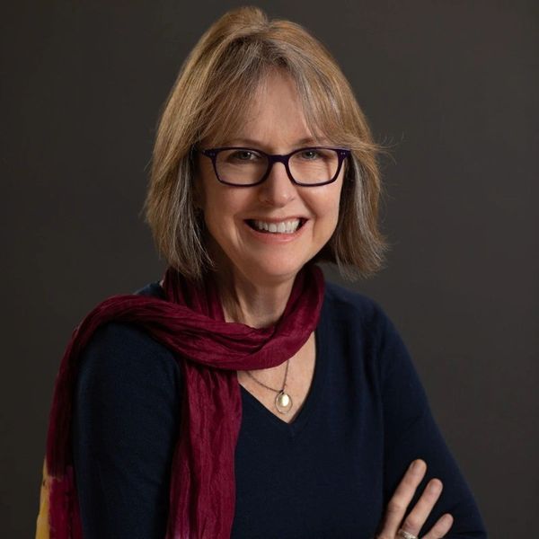 Smiling woman with glasses and a burgundy scarf poses against a dark background.