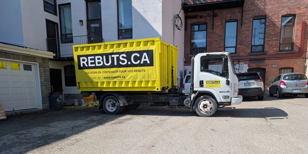 Yellow REBUTS.CA container truck parked in an urban lot beside buildings.