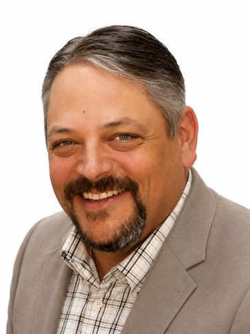 Smiling middle-aged man in a beige blazer and checkered shirt against a white background.