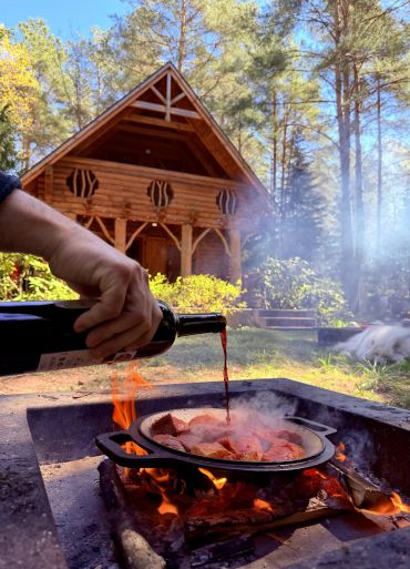 Pouring wine into a pan over an open fire in front of a wooden cabin.