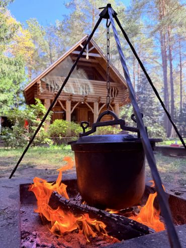 Cast iron pot cooking over open fire near a wooden cabin in the woods.
