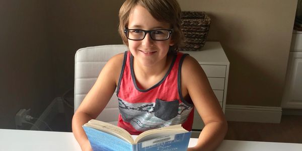 A young boy with glasses reading a book at a white table.
