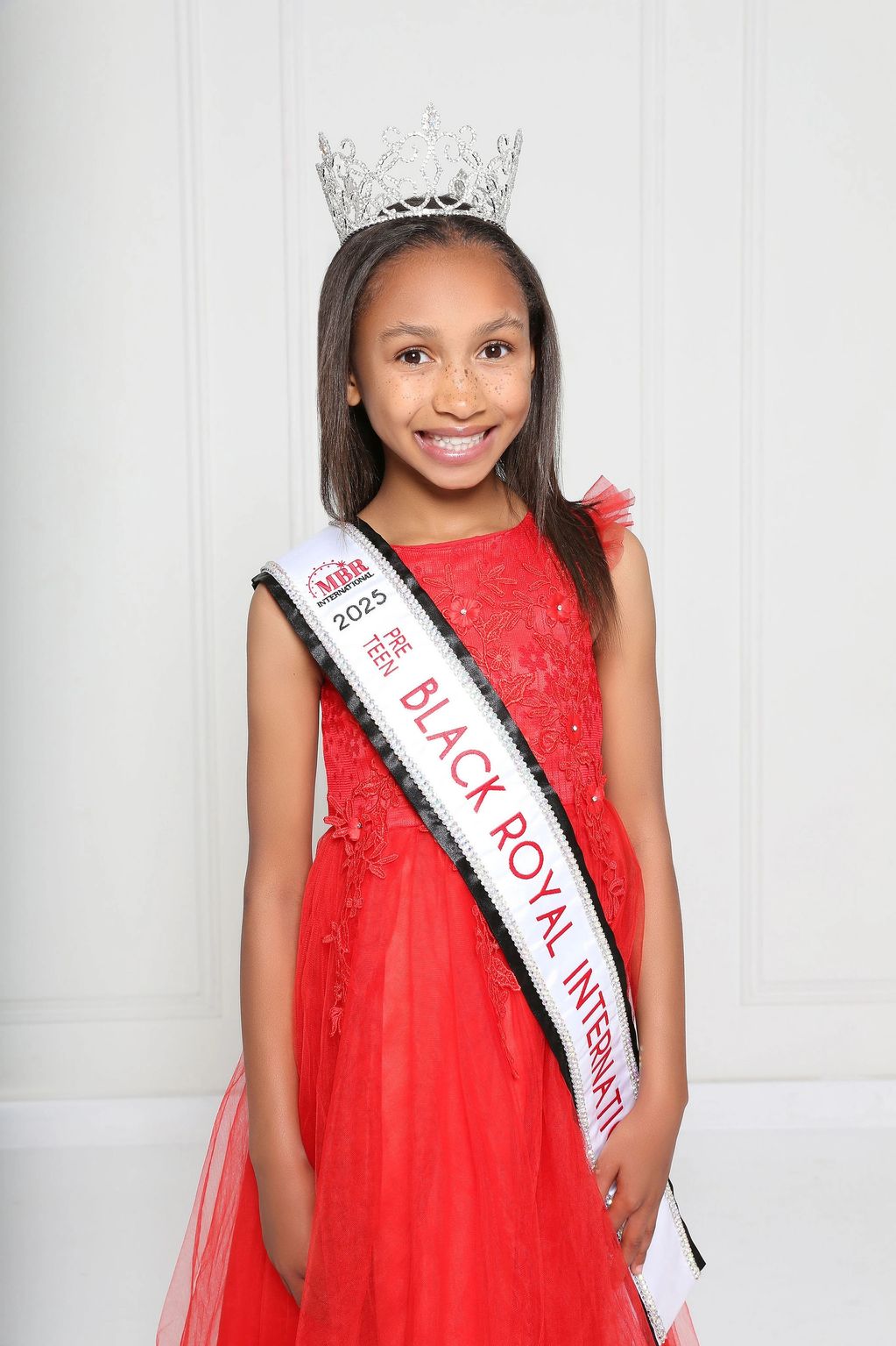 Young girl wearing a crown and sash, smiling in a red dress.