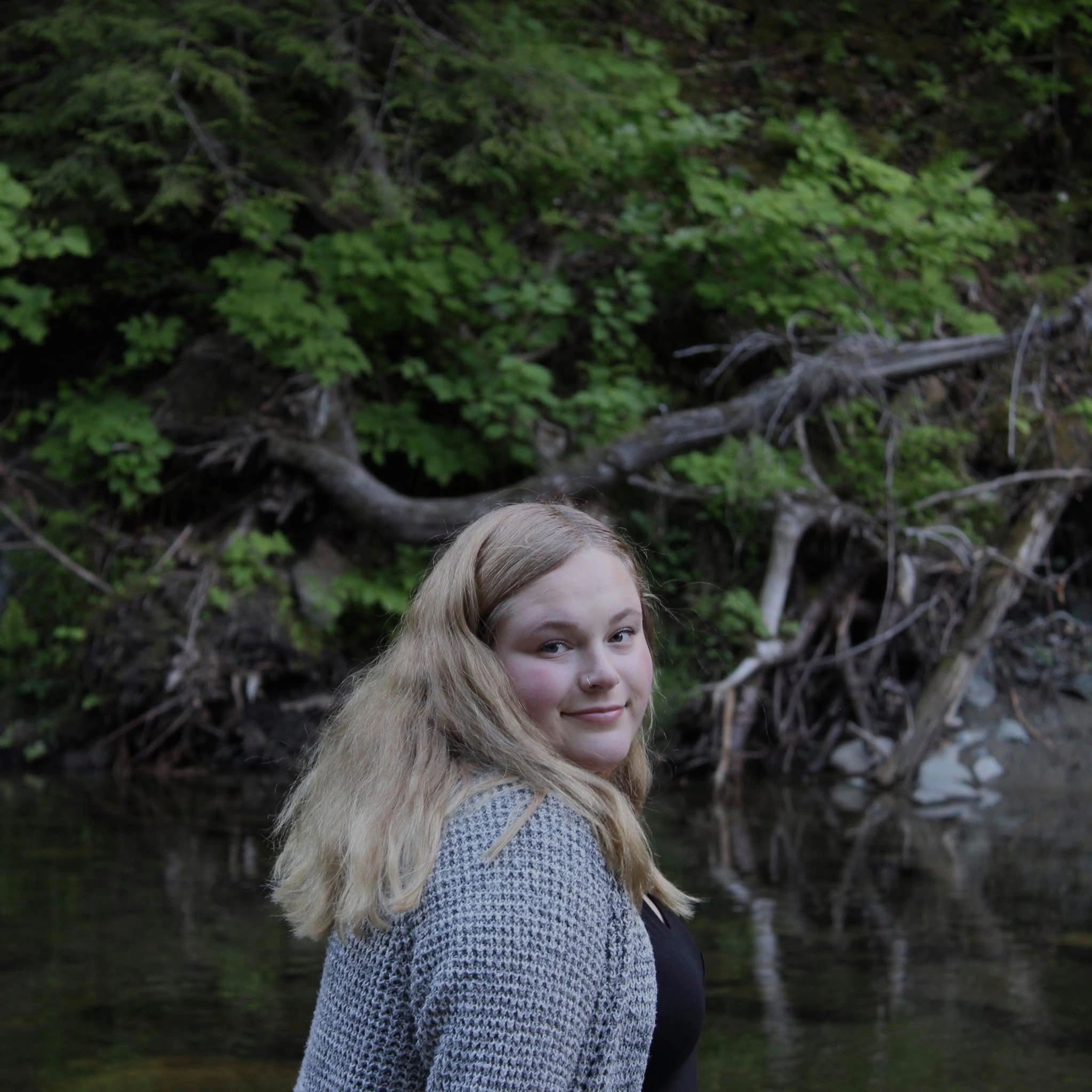 Young woman with long blonde hair smiling outdoors near greenery and water.