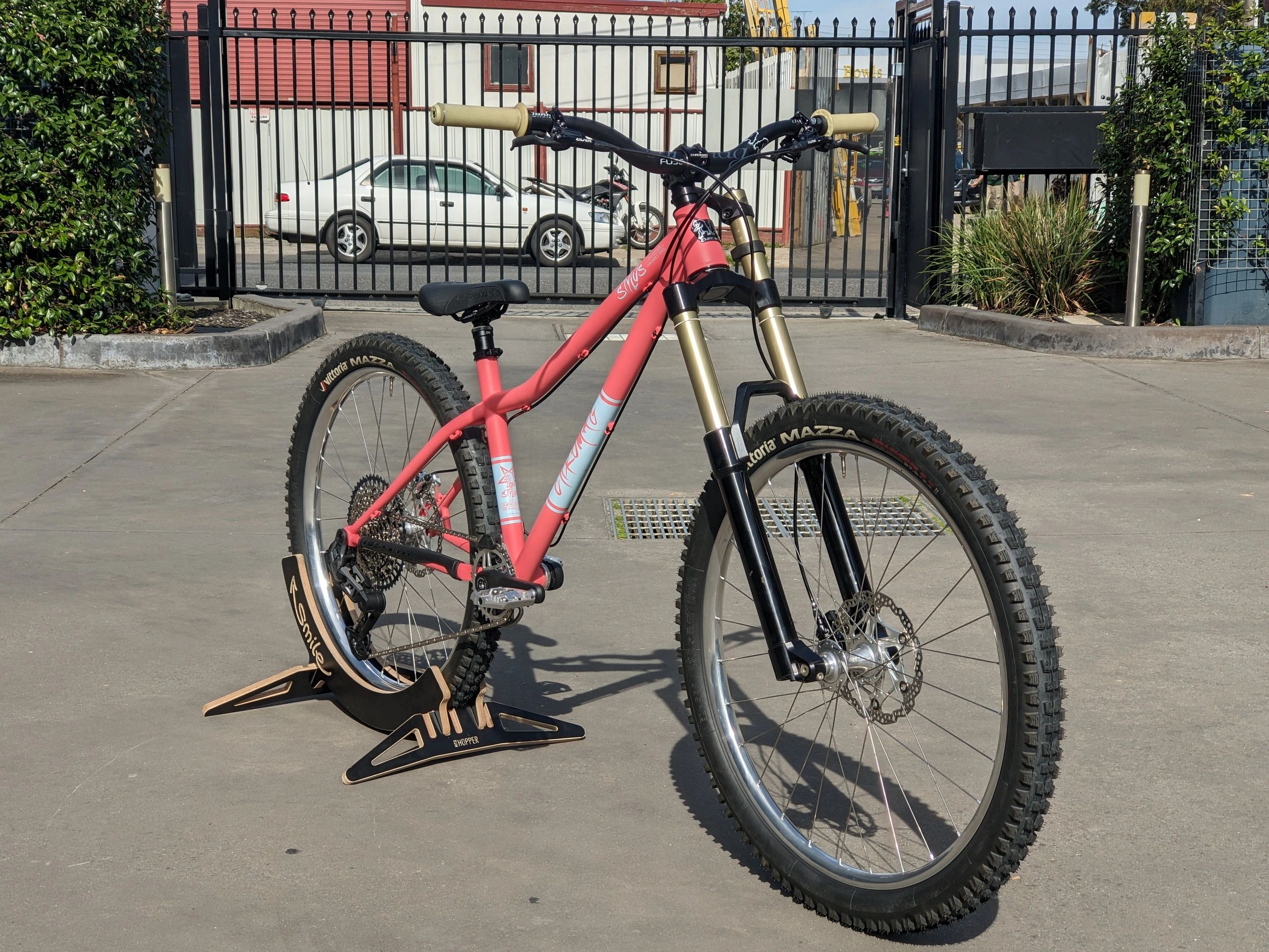A pink and black mountain bike on a stand outdoors with a gate and car behind.