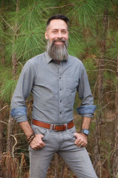 Man with a long beard standing outdoors in front of pine trees.