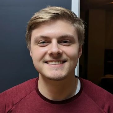 Young man with blond hair smiling in a casual setting.