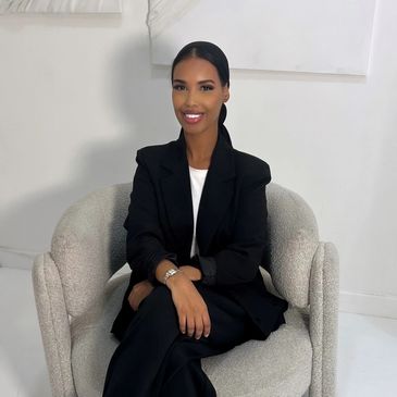 A smiling woman in a black blazer sitting on a modern beige chair.